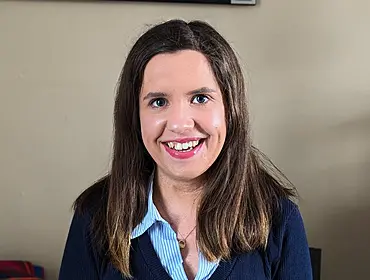 A woman with long brown hair smiles at the camera while sitting in front of a beige wall.