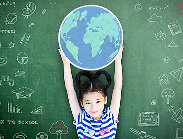 A small girl holds a cutout of the globe, against a blackboard with STEM signs on it.