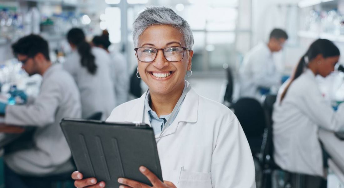 An older woman of colour holds a device and stands in front of a full lab.