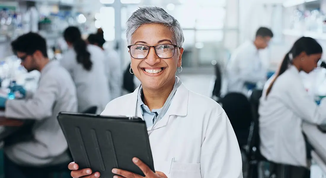 An older woman of colour holds a device and stands in front of a full lab.