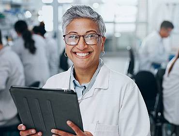 An older woman of colour holds a device and stands in front of a full lab.