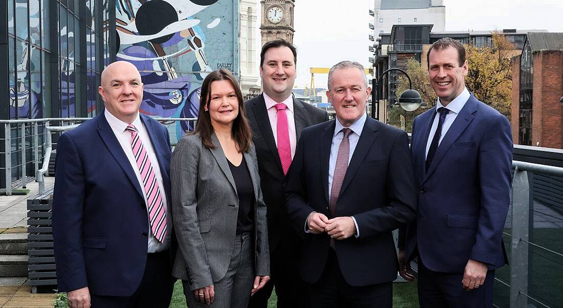 Five people in suits standing outdoors with a tall clock tower in the back.