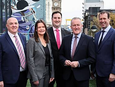 Five people in suits standing outdoors with a tall clock tower in the back.