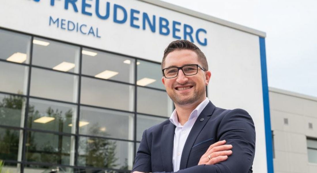 A man dressed in a suit and wearing glasses smiles and crosses his arms confidently outside of a building with the words ‘Freudenberg Medical’ on it.