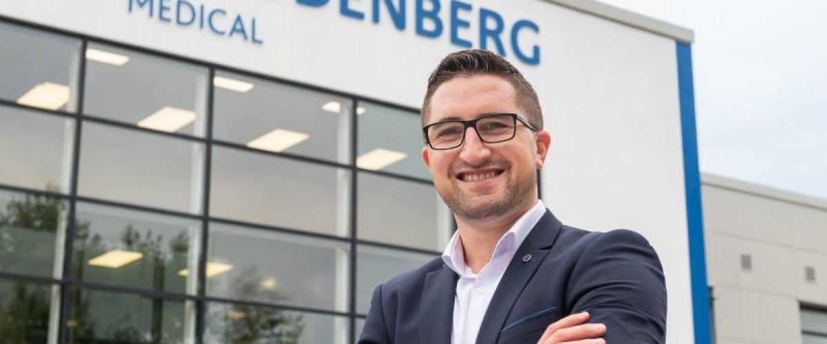 A man dressed in a suit and wearing glasses smiles and crosses his arms confidently outside of a building with the words ‘Freudenberg Medical’ on it.