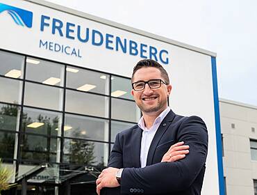 A man dressed in a suit and wearing glasses smiles and crosses his arms confidently outside of a building with the words ‘Freudenberg Medical’ on it.