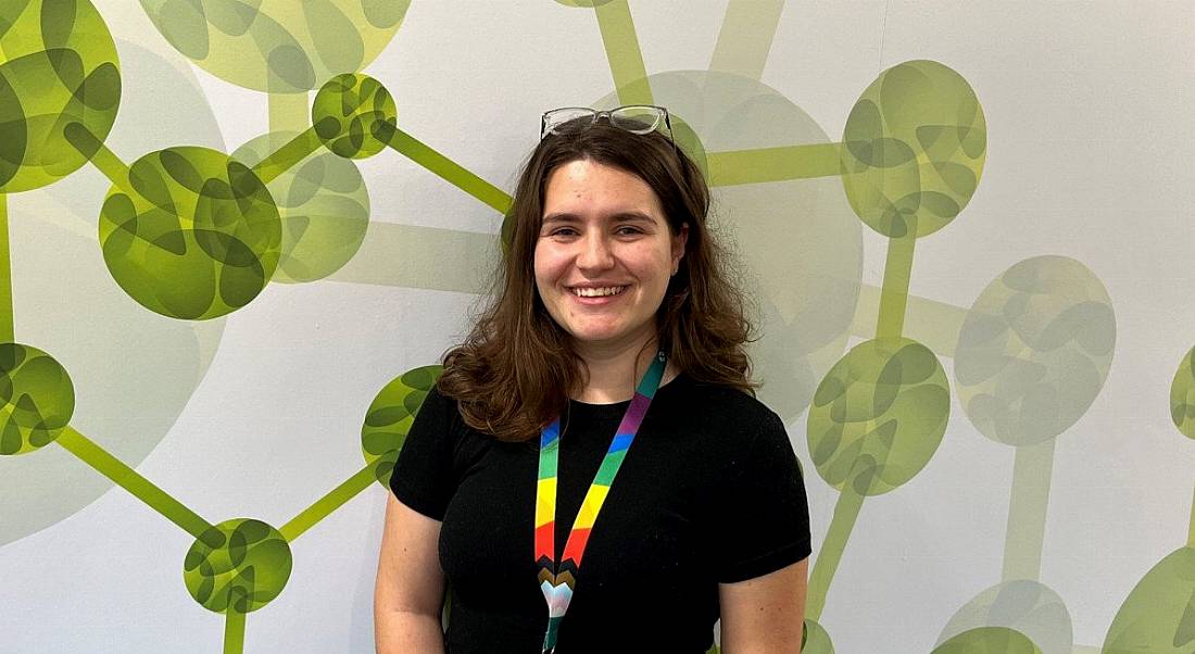 A woman wearing a black T-shirt with a rainbow-coloured lanyard smiles while standing in front of a wall with a green pattern on it.