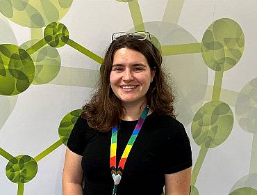 A woman wearing a black T-shirt with a rainbow-coloured lanyard smiles while standing in front of a wall with a green pattern on it.