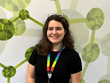 A woman wearing a black T-shirt with a rainbow-coloured lanyard smiles while standing in front of a wall with a green pattern on it.