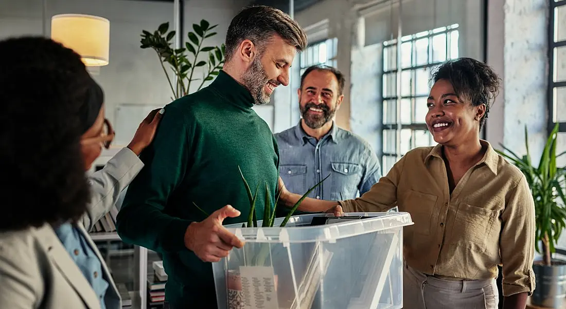 A man holding a box of his work possessions is greeted by his new co-workers.