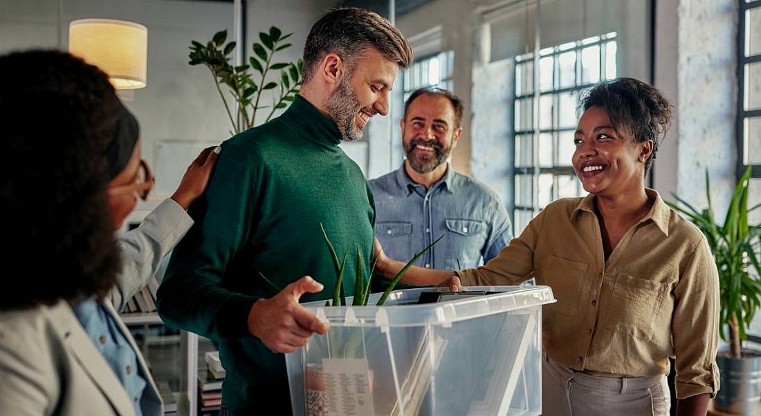 A man holding a box of his work possessions is greeted by his new co-workers.