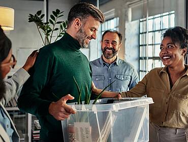 A man holding a box of his work possessions is greeted by his new co-workers.