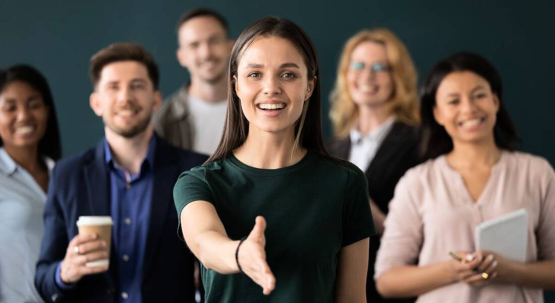 A woman in front of a team extends her hand to welcome you to a new career.