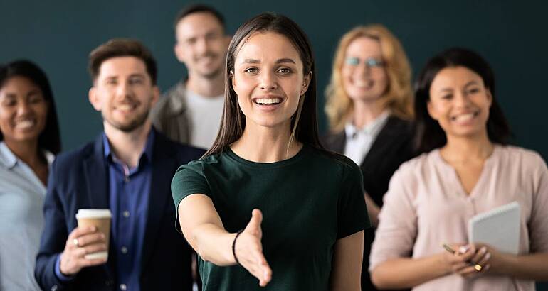 A woman in front of a team extends her hand to welcome you to a new career.