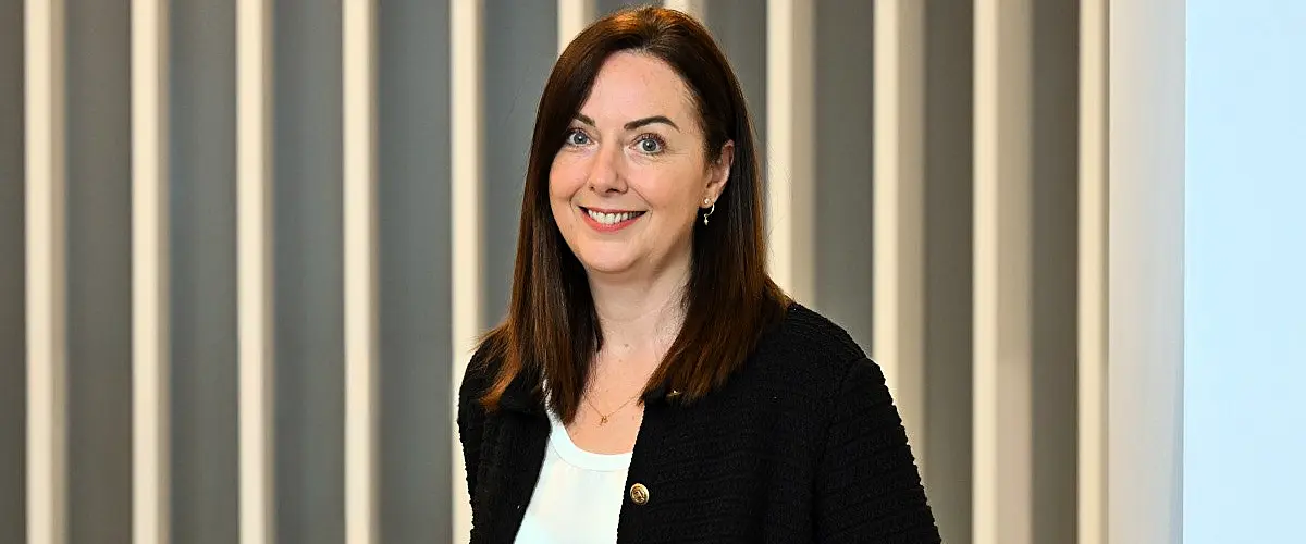 A woman wearing a black blazer stands in an office smiling at the camera.