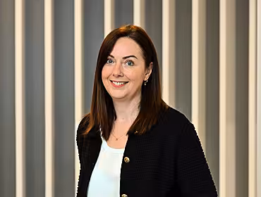 A woman wearing a black blazer stands in an office smiling at the camera.
