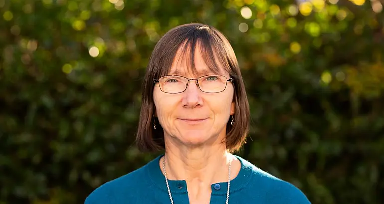 A woman with shoulder-length brown hair and glasses smiles at the camera in front of shrubbery.