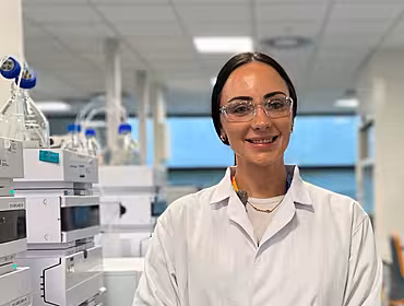 A woman with dark hair stands in a laboratory wearing a white lab coat and clear safety goggles.