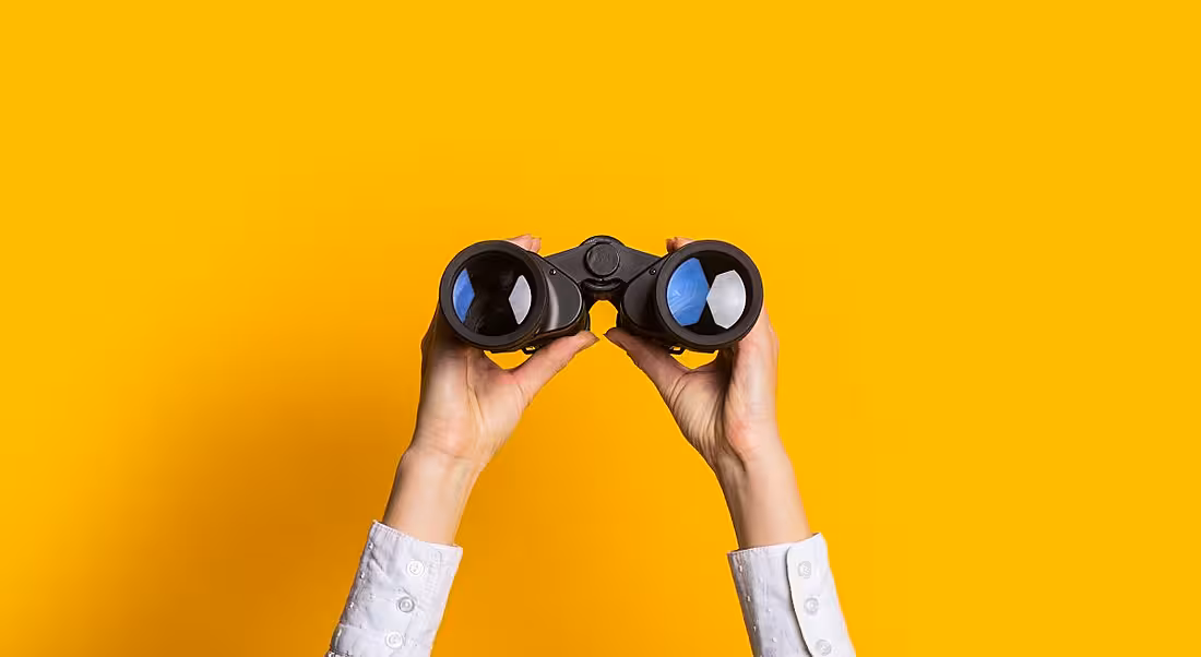 A yellow background, with a pair of hands holding up binoculars.