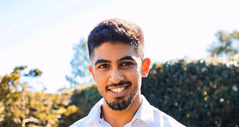 A headshot of a young man with dark hair wearing a shirt in an outdoor sunny setting.