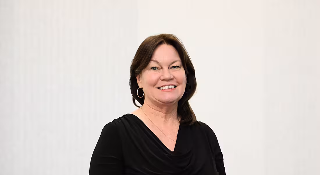 A headshot of a woman with dark hair against a grey wall smiles at the camera.