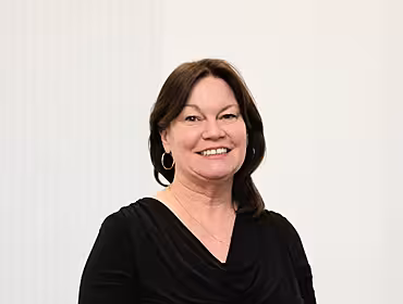 A headshot of a woman with dark hair against a grey wall smiles at the camera.