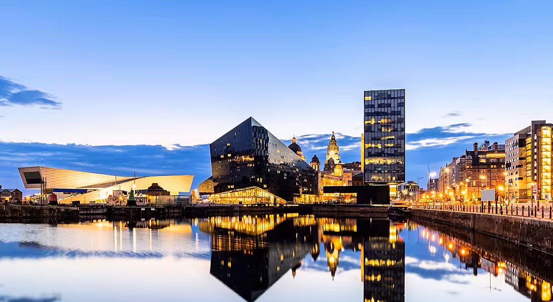 The Liverpool skyline pier at sunset. Brightly lit buildings line the edge of still water with perfect reflections in the water.