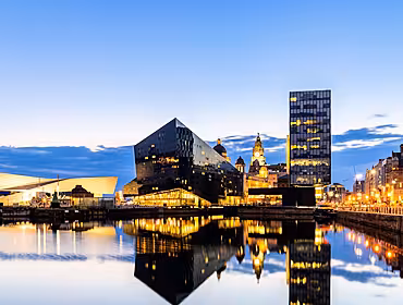 The Liverpool skyline pier at sunset. Brightly lit buildings line the edge of still water with perfect reflections in the water.