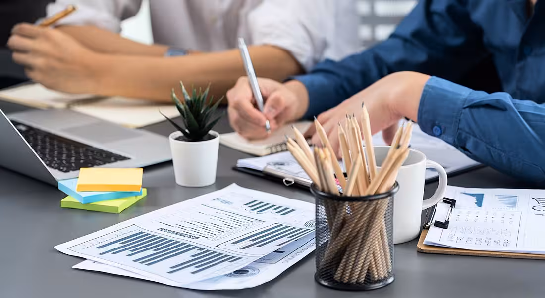 Two employees surrounded by charts, pens, paper and tech, representing the workplace.