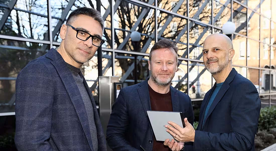 Three men in blazers stand in a line smiling at the camera in an outdoor glass house. One is holding a digital tablet.