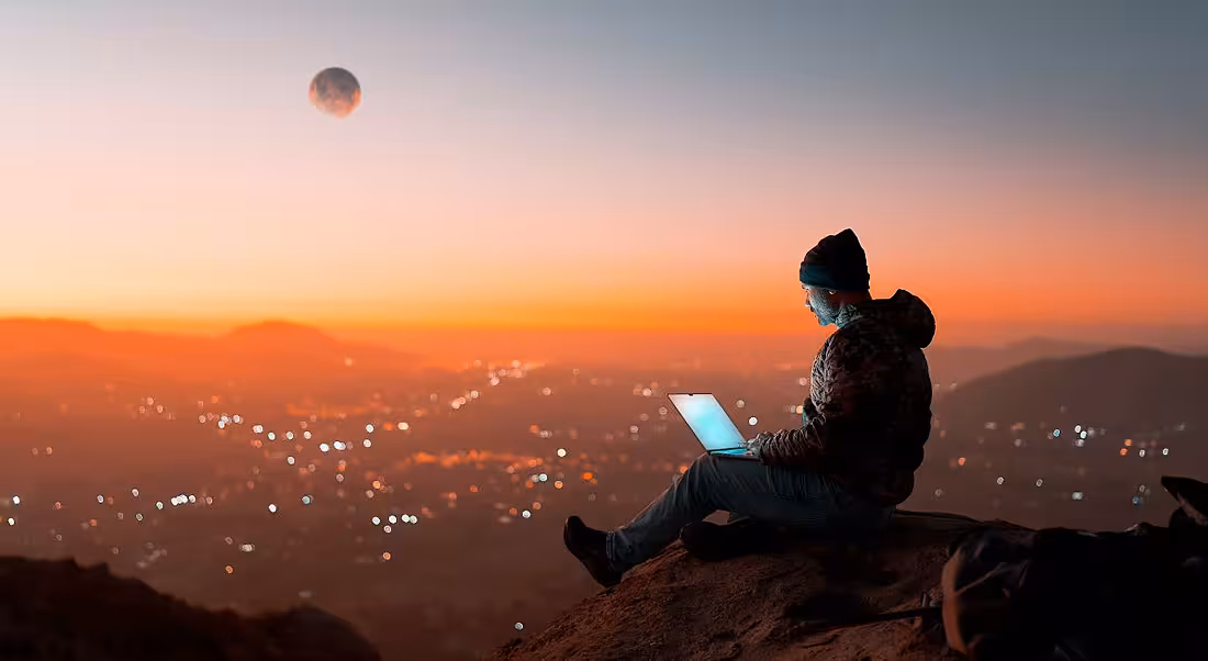 A man on a laptop works as a digital nomad, with a sunset and mountaintop view in front of him.