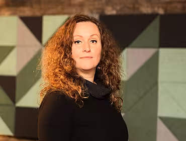 A woman with curly brown hair wears a black jumper and faces the camera.