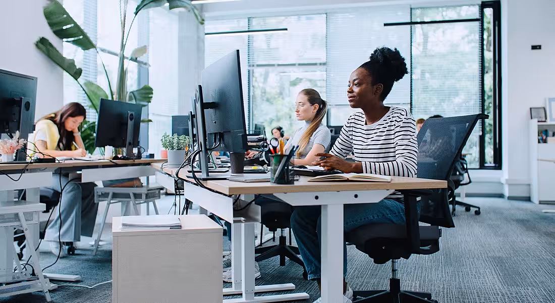 A young woman in casual clothes sits at her work computer, surrounded by her co-workers.