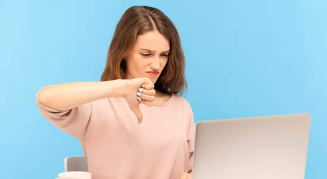 Against a blue background a woman in pink points her thumb down in front of a laptop.