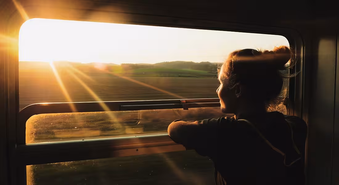 A young woman on a train looking out a window into the sunset.