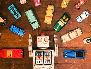 An aerial image of a toy robot lying on a wooden floor. It is surrounded by toy cars to symbolise driving the AI revolution.