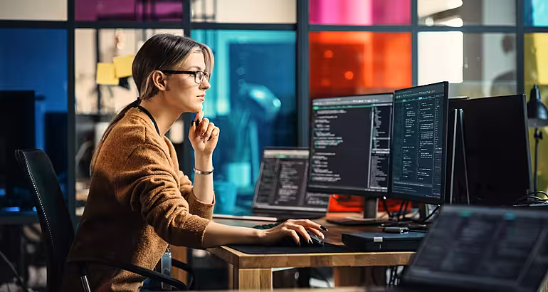 A woman sits at a desk with multiple computer screens showing lines of code.