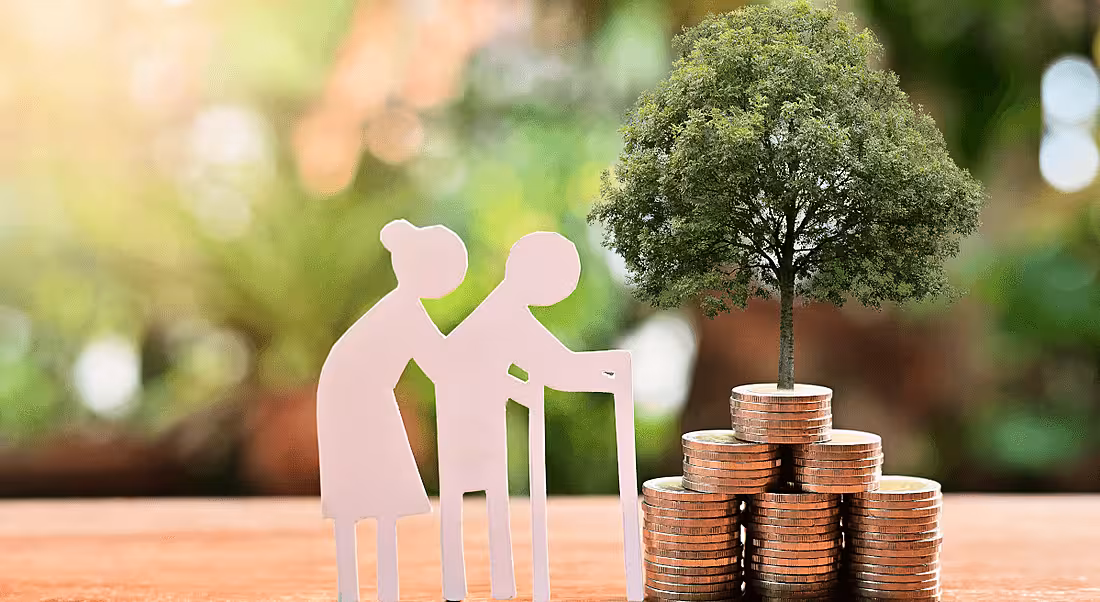Two cutouts of elderly people walk towards a tree growing out of a stack of coins, representing delayed retirement.