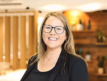 A headshot of a woman with blonde hair and glasses smiling at the camera in a bright office space.