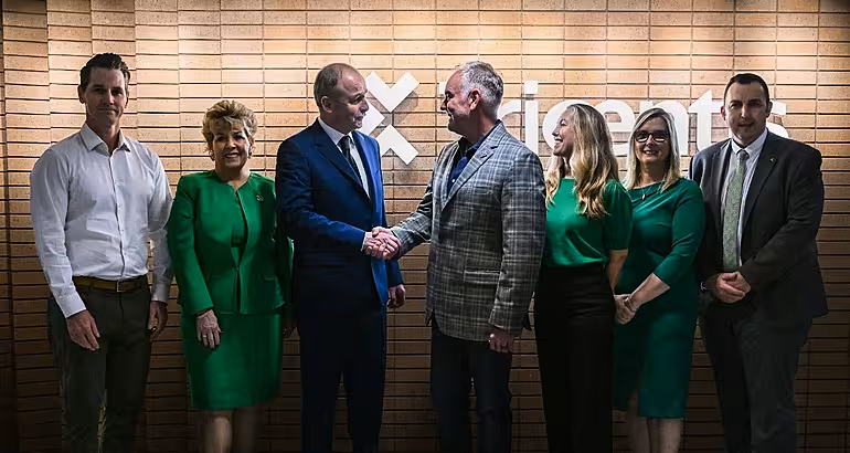 A group of men and women stand in a line in front of the Tricentis logo on a brick wall. Taoiseach Micheál Martin is in the centre shaking hands with another man.
