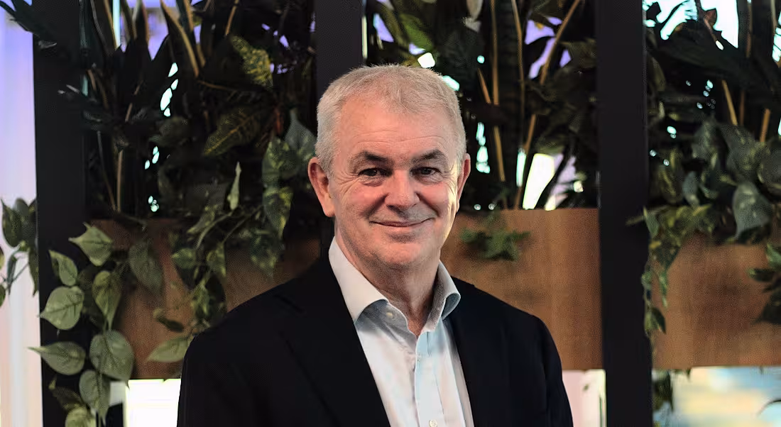 A man wearing a dark blazer and open-collar shirt stands in front of some greenery in a brightly lit space.