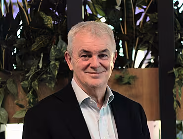 A man wearing a dark blazer and open-collar shirt stands in front of some greenery in a brightly lit space.