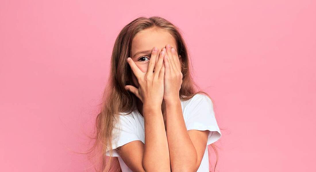 Little girl covering her face with her hands against pink background.