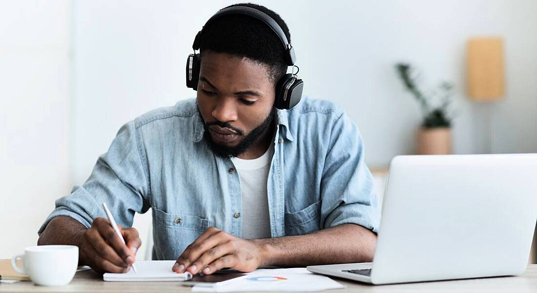 A man in casual clothes works at a desk with a pair of headphones on and a laptop in front of him.