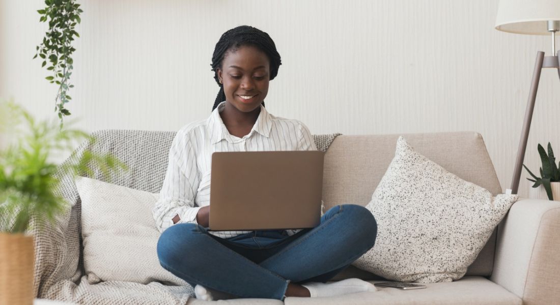 A woman is sitting on her couch at home with her laptop, working remotely.