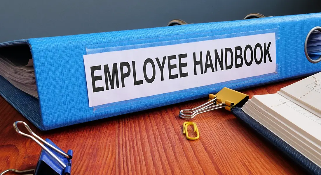 A blue employee handbook to onboard new staff on a wooden office desk.