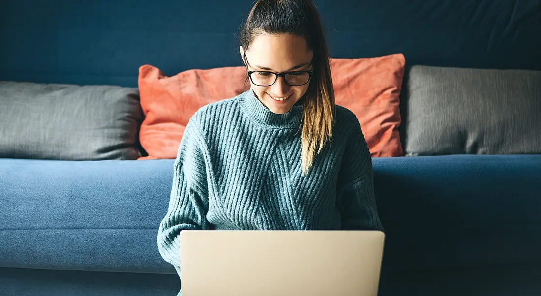 A woman in casual clothes is working remotely on a laptop in front of a navy couch.