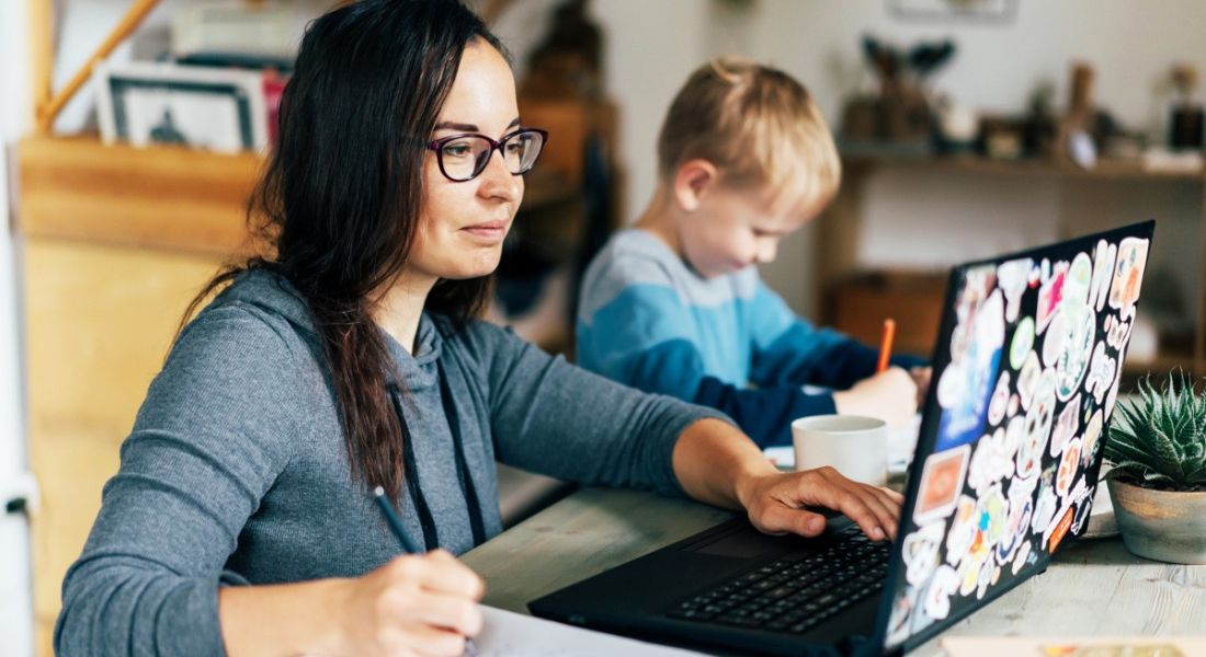 A woman is working at home on her laptop beside her child doing homework in their kitchen.