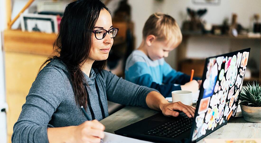 A woman is working at home on her laptop beside her child doing homework in their kitchen.