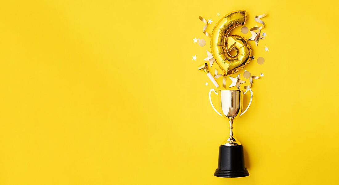 Gold foil number six balloon exploding, suspended over a trophy surrounded by confetti and streamers, on a bright yellow background.